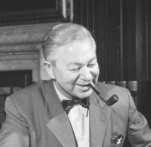 A black-and-white image of Arne Jacobsen, sitting and smiling, looking down and with a pipe in his mouth.