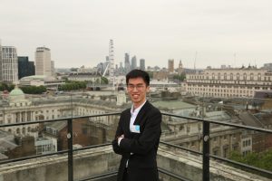A student, smiling, with his arms crossed and with London skyline in the background.