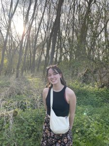 A photo of a white woman smiling at the camera, stood amongst woodland.